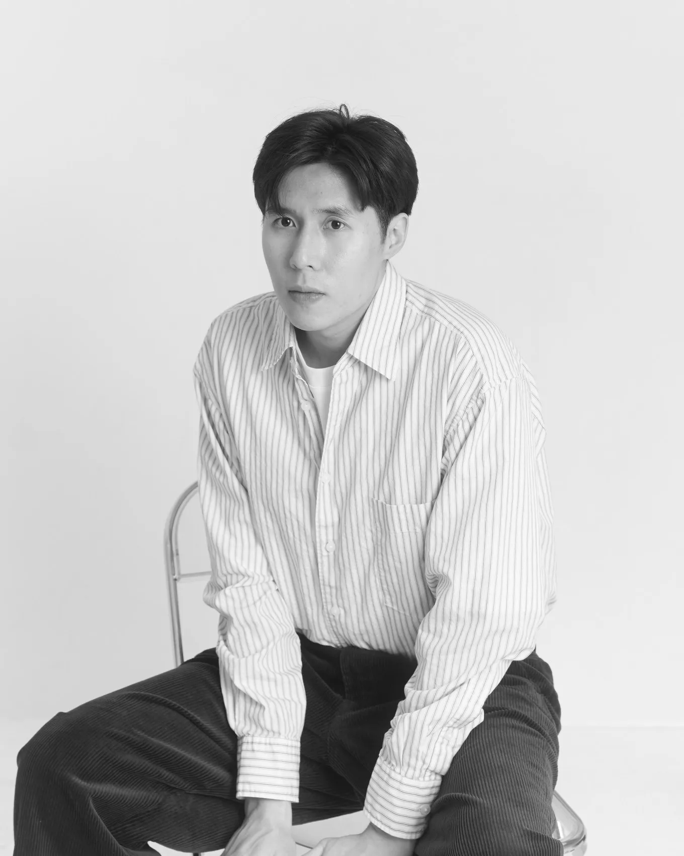Derick Tsang seated on a stool in a striped shirt and corduroy trousers, looking at the camera.